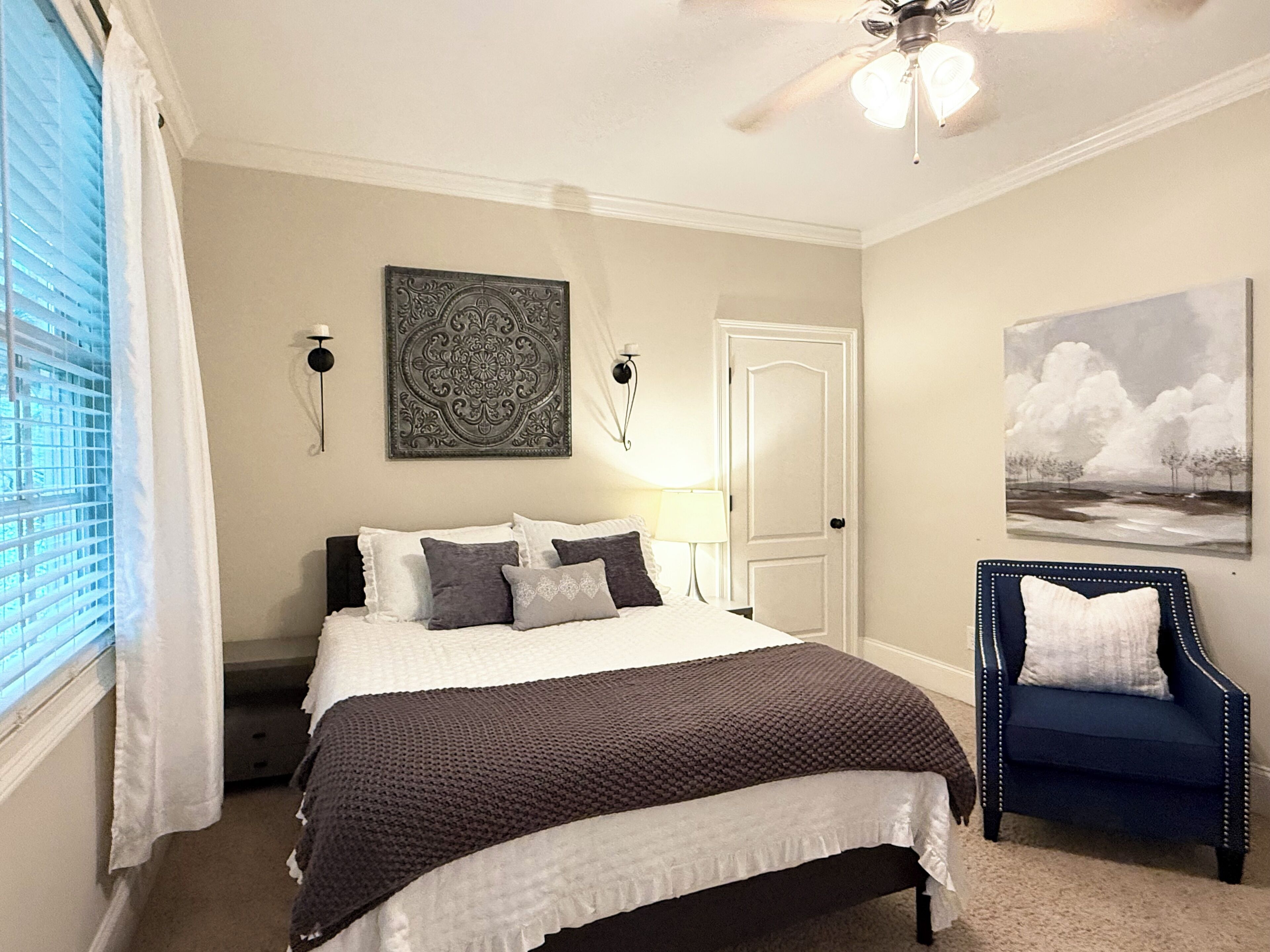 Quarto com cama, cadeira, ventoinha no teto, porta e pintura na parede.