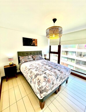 A bedroom with a bed, a nightstand, a lamp, a hanging light fixture, a window, and a view of buildings outside. - Las Condes Apt | Prime Location + Fully Equipped (Las Condes)