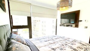 A bedroom with a bed, a television, a window, and a view of buildings. - Las Condes Apt | Prime Location + Fully Equipped (Las Condes)
