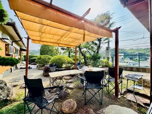 An outdoor patio area with a covered seating arrangement, including a table and chairs, surrounded by rocks and greenery. - Fumiko-no-tei Residence with Ocean View; accommodates up to 17–34 people. Garden & Barbecue (西牟婁郡白浜町)
