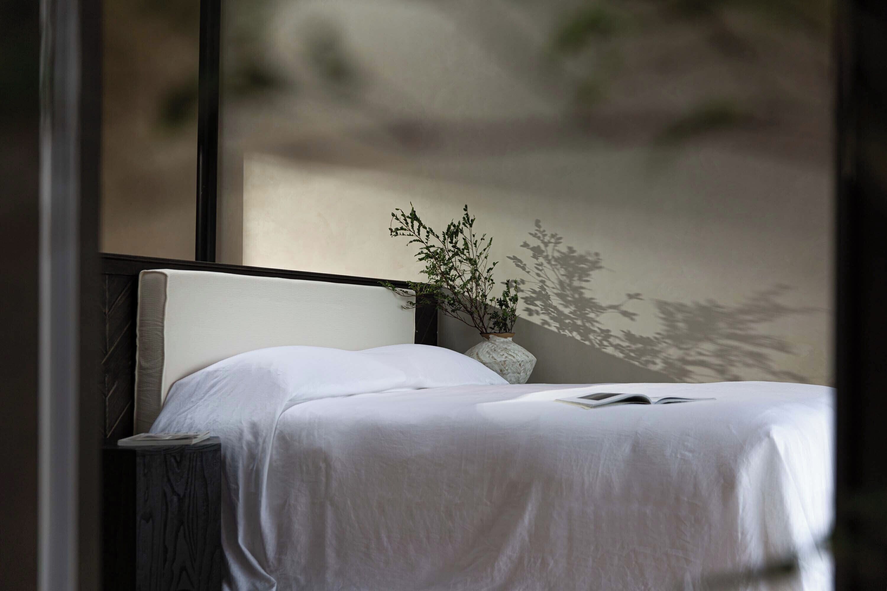 A neatly made bed with a white sheet, a small potted plant on the nightstand, and a window with a view of a building.