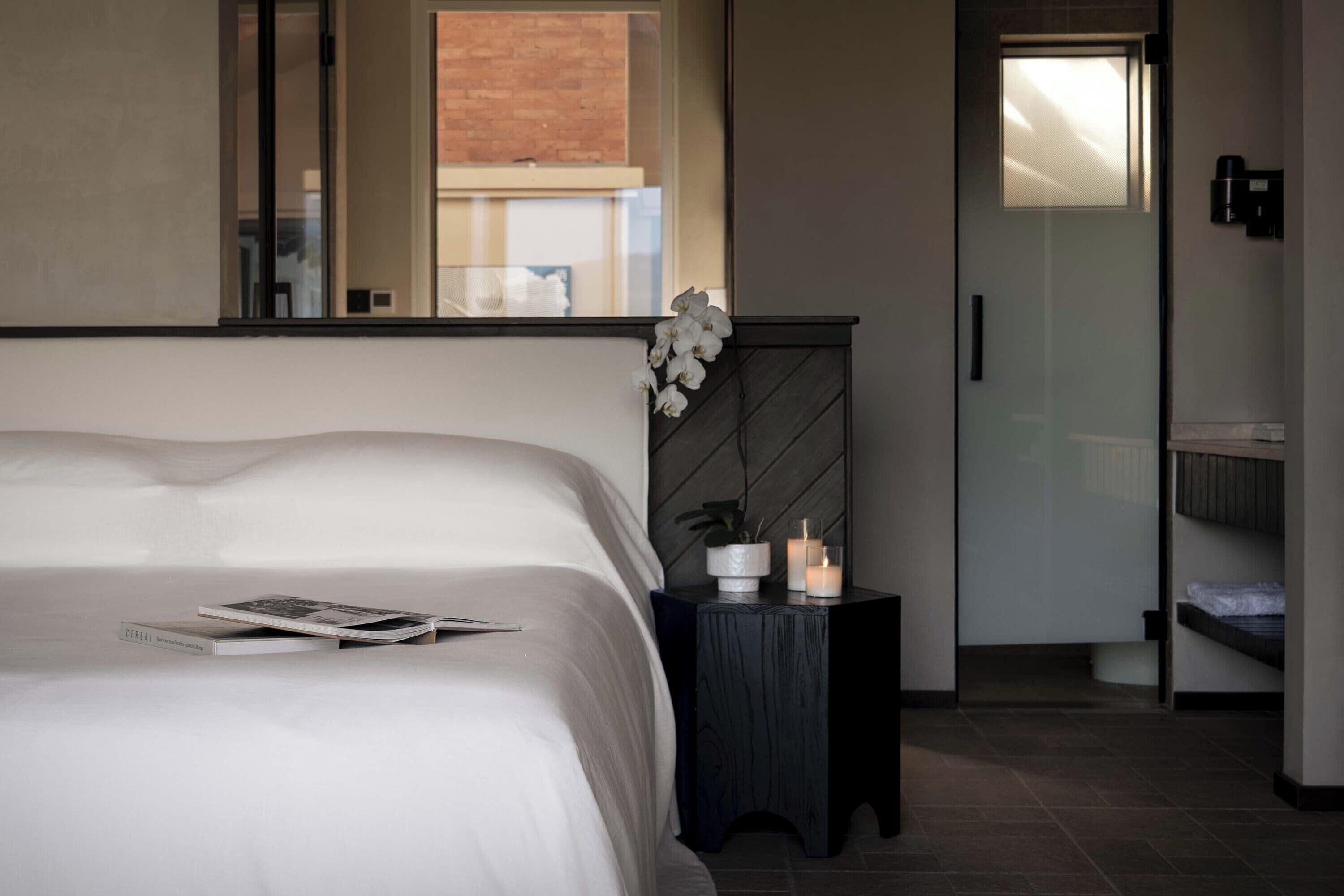 A modern bedroom with a large bed, a small side table with a plant and candles, a mirror, and a view of the outside.