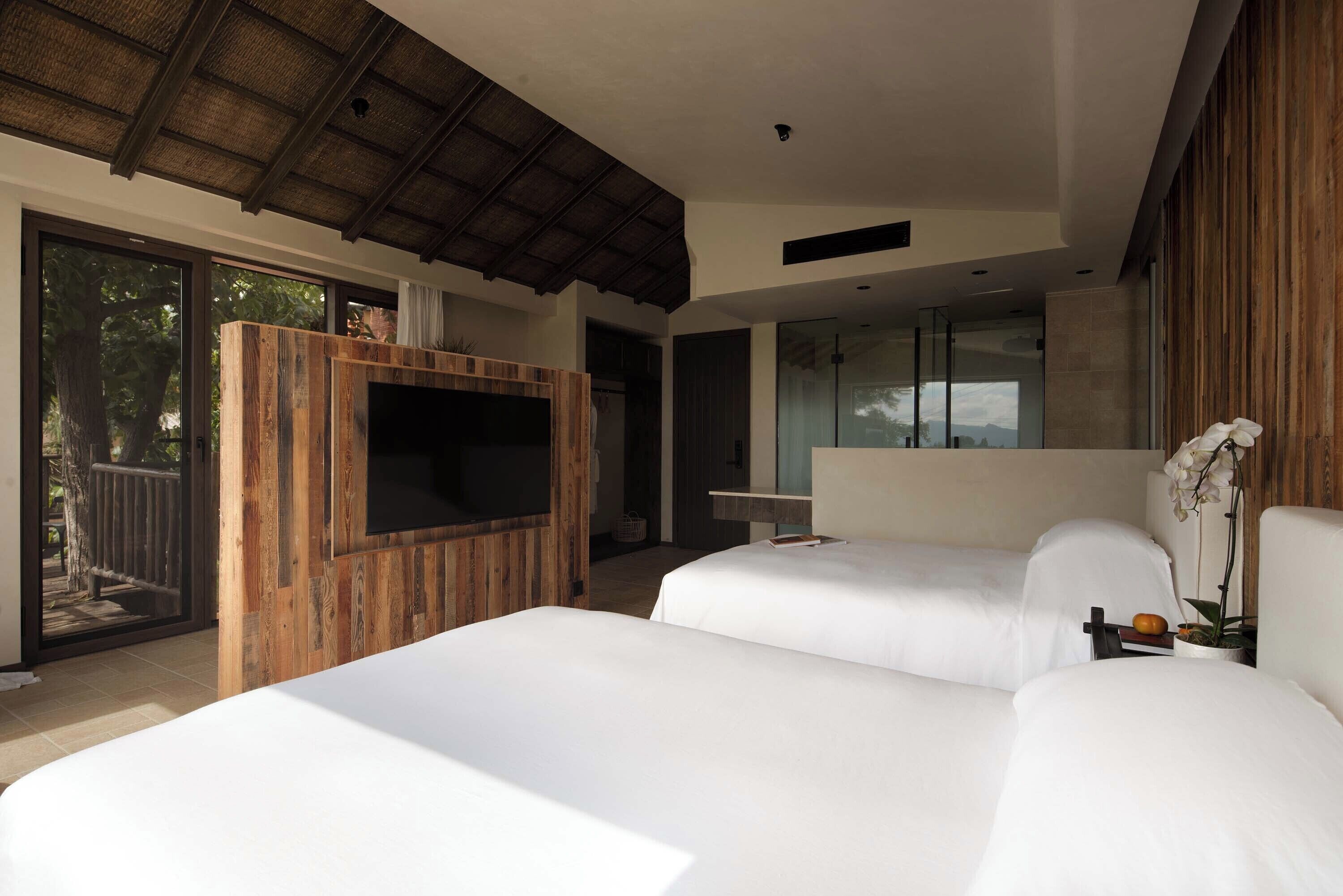 A modern bedroom with two beds, a wooden TV stand, and a large window overlooking a balcony.