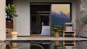 A modern hotel room with a large sliding glass door leading to a mountain view, a small wooden table, a chair, and a potted plant. - Snow Mountain (Lijiang)