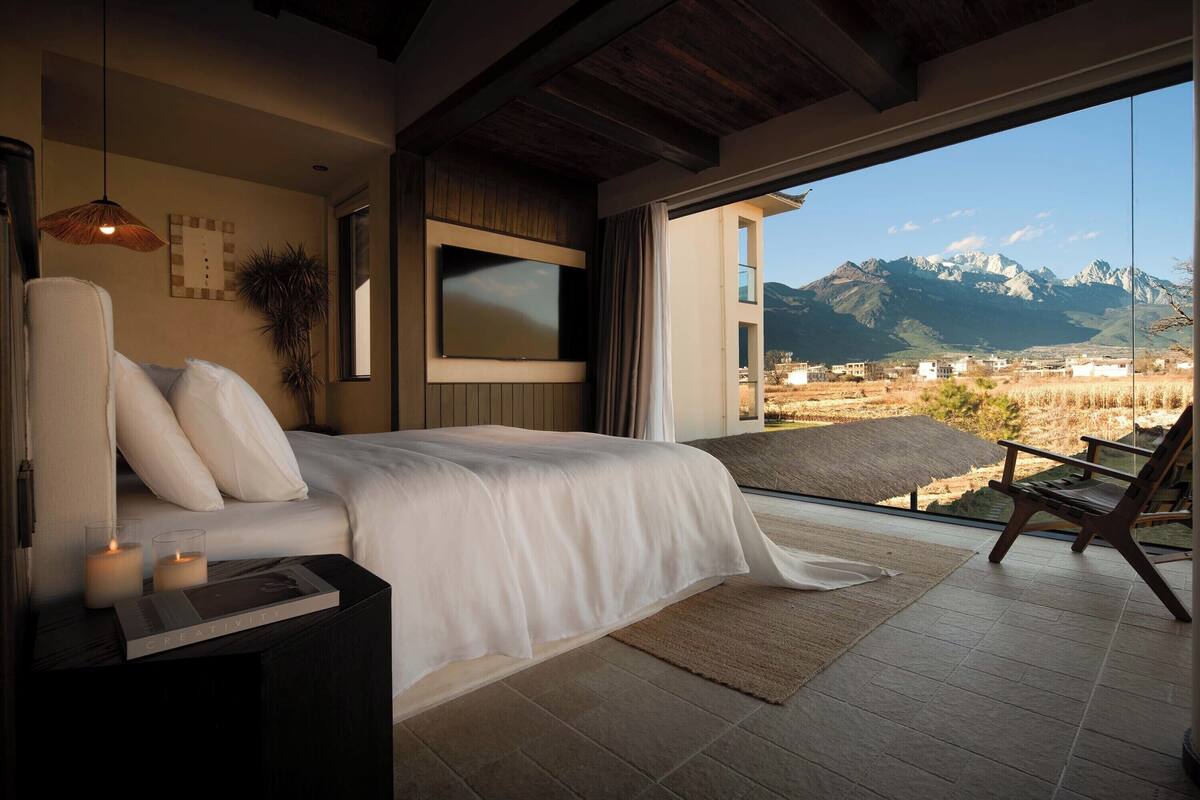 A bedroom with a bed, a TV, a window with a mountain view, and a chair.