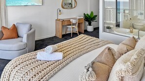 A modern hotel room with a bed, a desk, a chair, a large window with a view of the city, and a painting of a beach. - Luxury Oracle Tower 2 Ocean Views 22nd floor (Broadbeach)