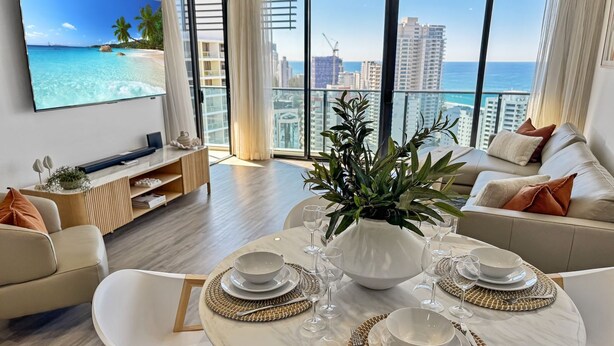 A modern living room with a dining area, large windows with a city view, and a flat-screen TV on the wall. - Luxury Oracle Tower 2 Ocean Views 22nd floor (Broadbeach)