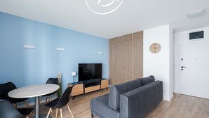 Un salon moderne avec un canapé gris, une table de salle à manger ronde entourée de chaises, un téléviseur posé sur un meuble en bois et une horloge murale.