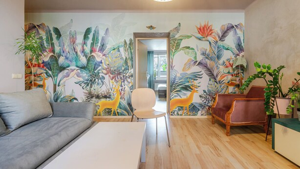 Ein Wohnzimmer mit einer großen Wandmalerei von tropischen Blättern und einem Reh, einem grauen Sofa, einem weißen Couchtisch und zwei Sesseln.