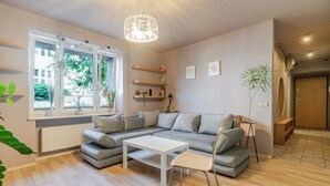 Ein modernes Wohnzimmer mit einer Eckcouch, einem weißen Couchtisch und einem Essbereich. Es gibt Regale mit Büchern und Dekoartikeln, eine Topfpflanze und ein großes Fenster mit Vorhängen.