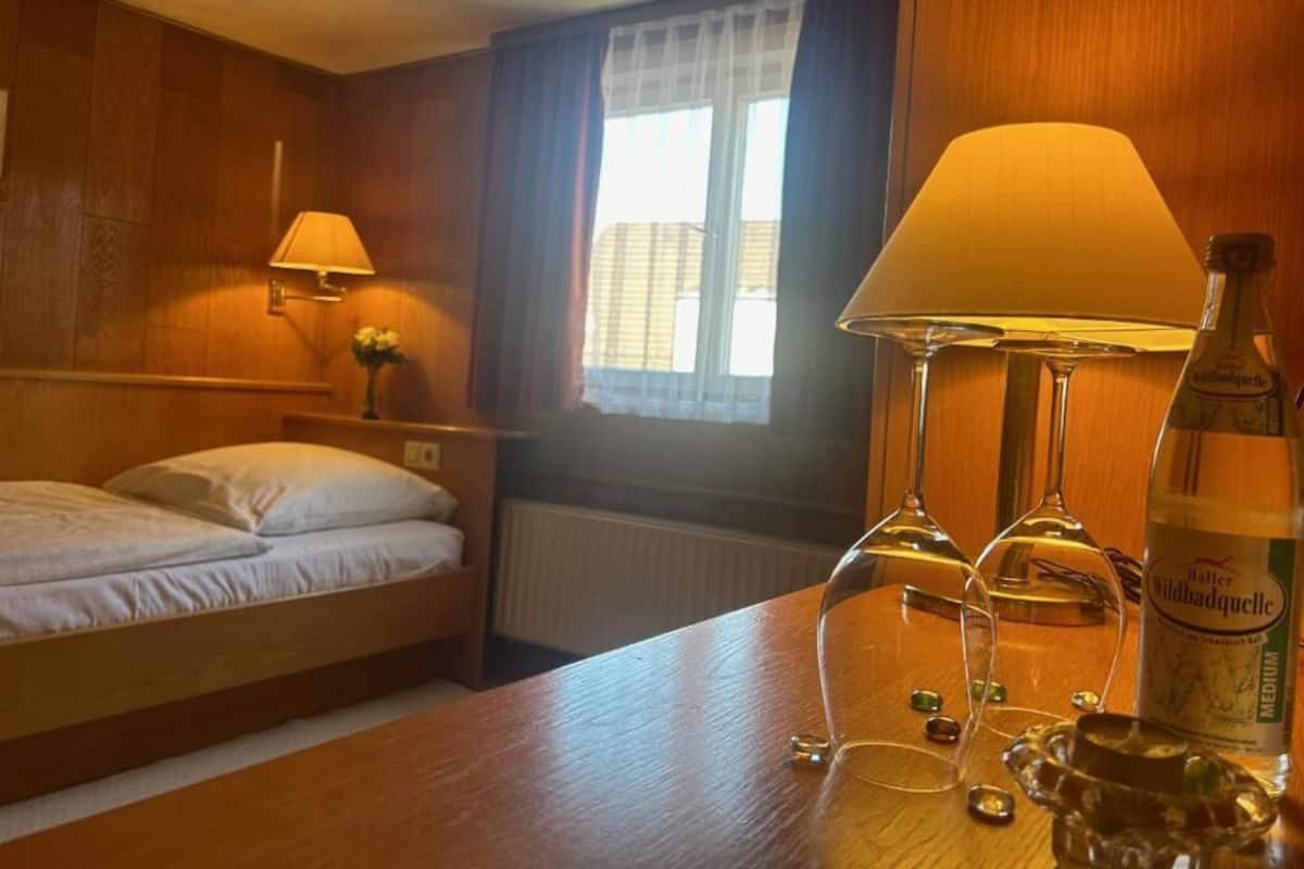 Een hotelkamer met een bed, een tafel met een fles Wildbadquell, twee wijnglazen en een lamp.