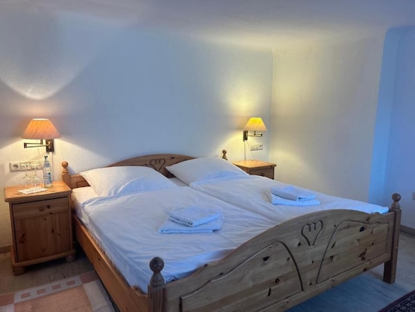 A neatly made bed with white linens and folded towels, two bedside lamps, and a wooden nightstand with a bottle and glass.