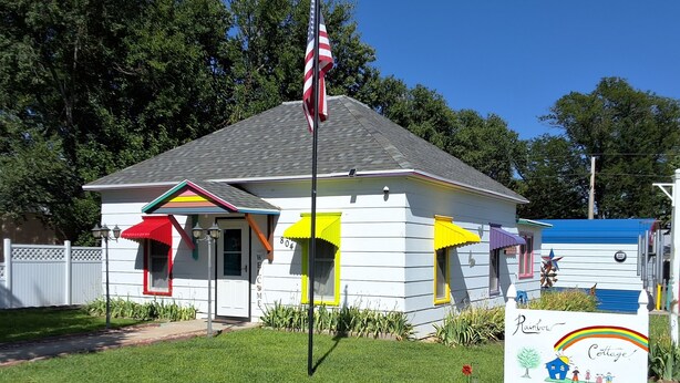 Una casetta colorata e accogliente, con un cartello di benvenuto, una bandiera e un'insegna che indica che si tratta del 'Rainbow Cottage'.