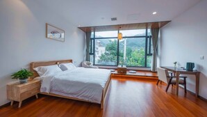 A modern bedroom with a large bed, a desk, and a view of greenery through large windows.