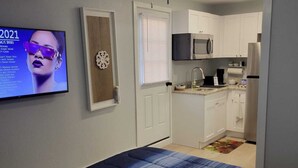 A bedroom with a bed, a ceiling fan, a TV, and a kitchenette in the background.