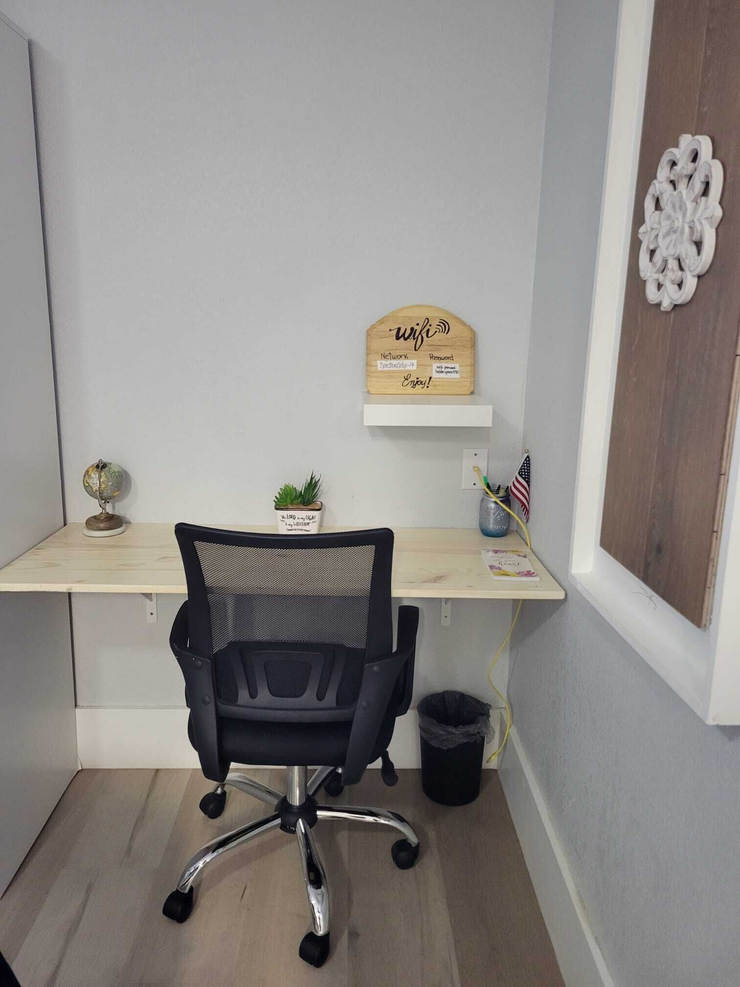 A small office space with a desk, a black office chair, a small potted plant, a wooden sign with 'wifi' and 'Enjoy!' on it, and a decorative wall piece.