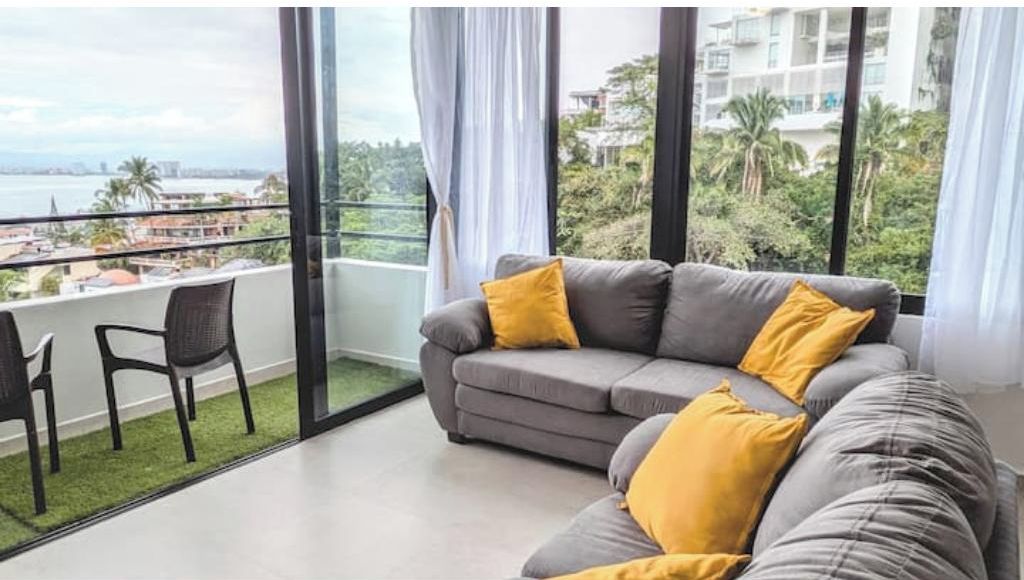 Ein modernes Wohnzimmer mit einem grauen Sofa und gelben Kissen, einem schwarzen Sessel und einem großen Fenster mit Blick auf Stadt und Grünflächen.