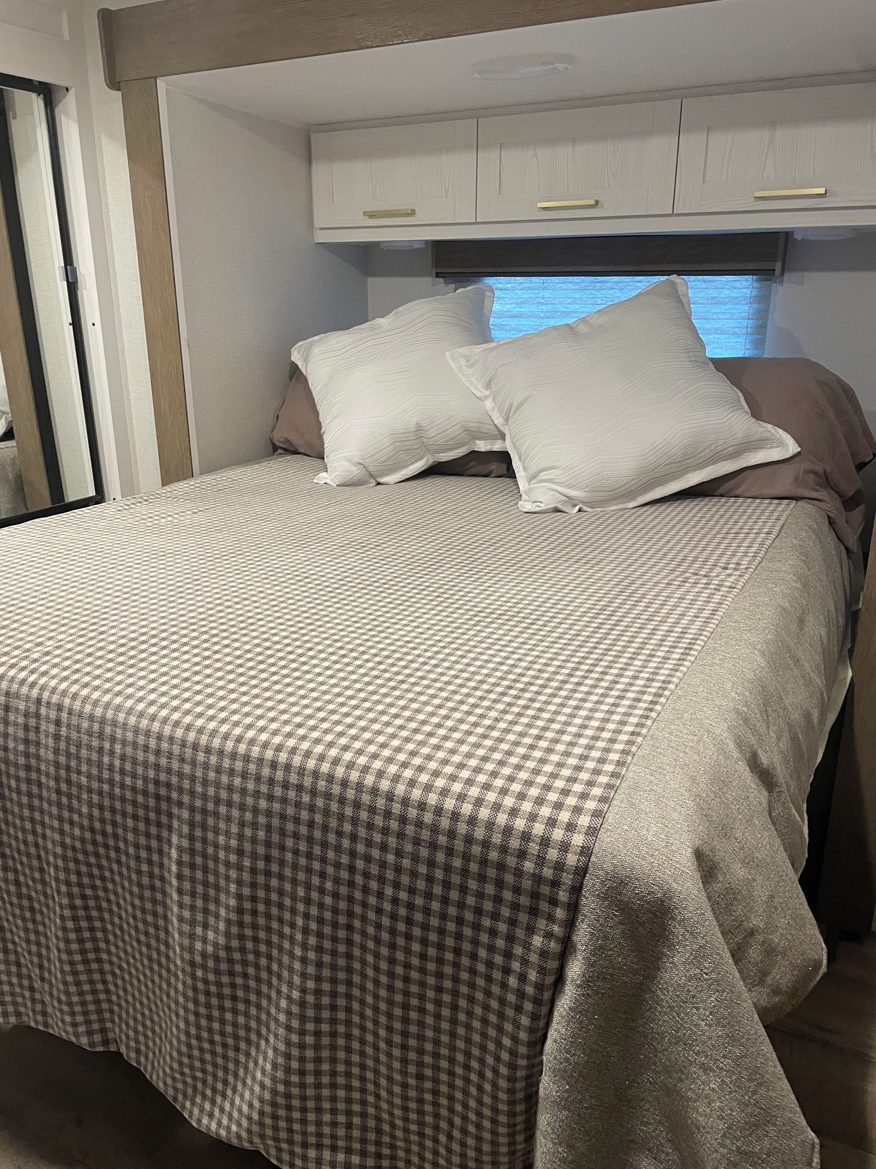 Een net opgemaakt bed met een geruit beddegoed en twee kussens, een raam met zonwering en een witte garderobe met gouden handgrepen.
