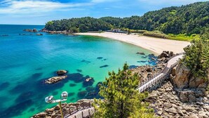 A coastal landscape with a sandy beach, clear turquoise waters, rocky outcrops, and a forested hill. - Centum Mark Kimstay Yangyang (Yangyang)