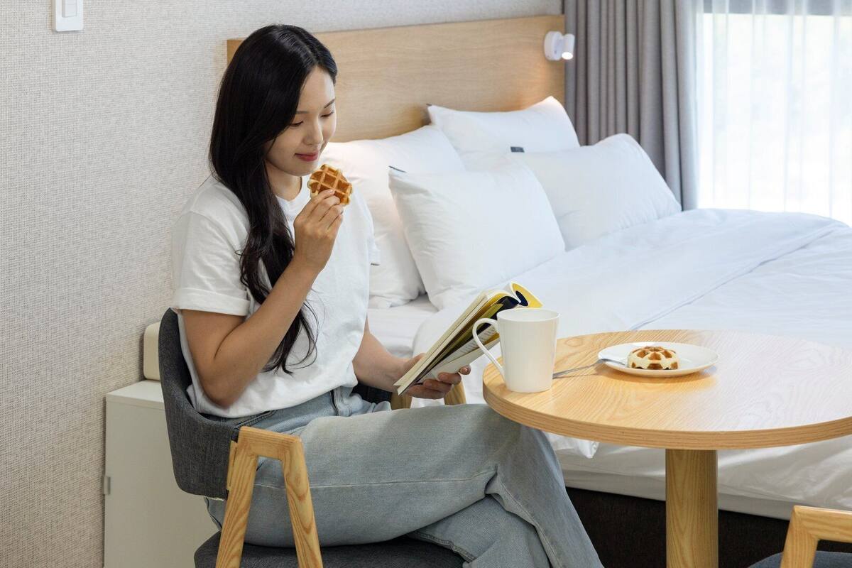 A person sitting at a round wooden table in a hotel room, eating a waffle and reading a book.