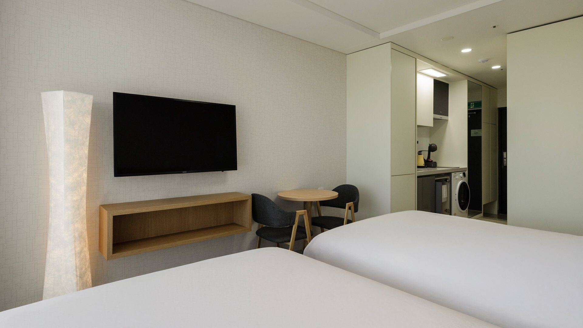 Habitación de hotel con dos camas, un televisor montado en la pared, una mesita con sillas y una lavadora en la esquina.