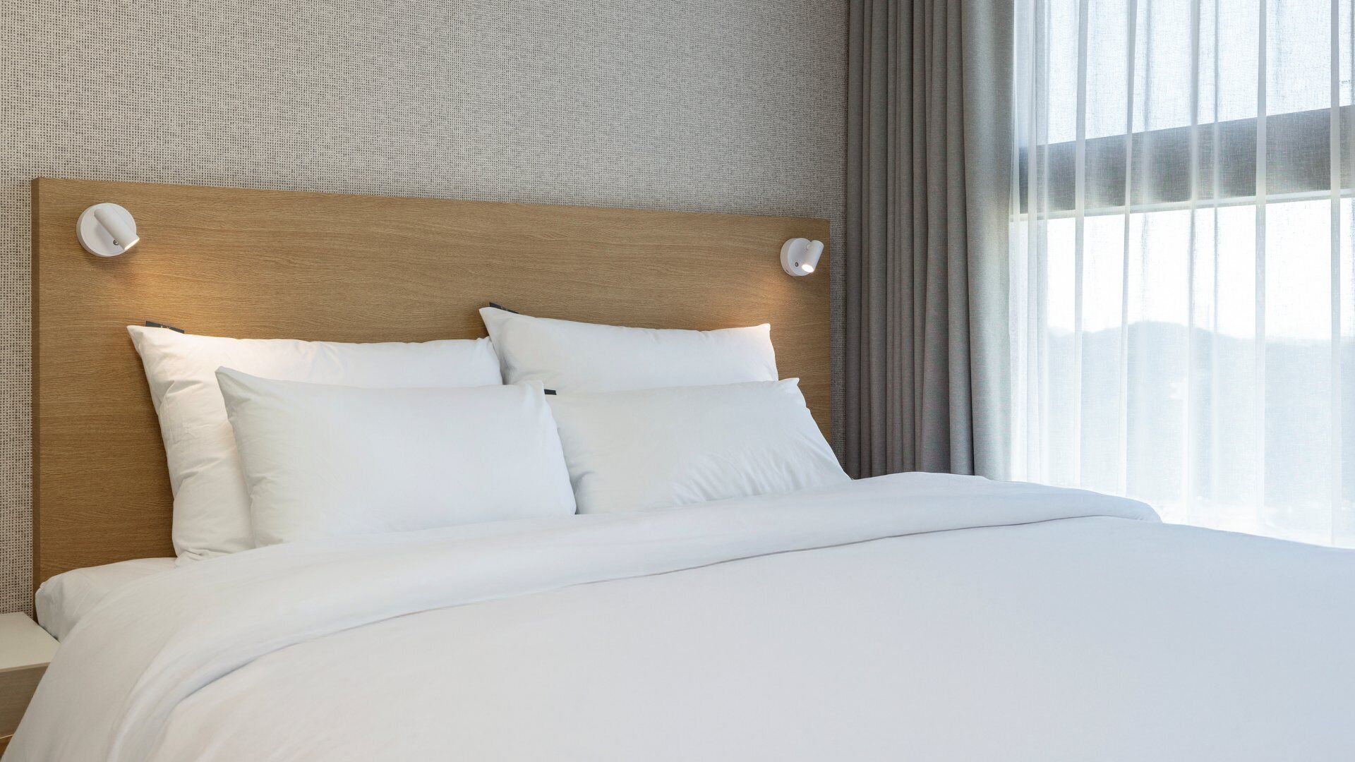 A hotel room with a wooden headboard, white bedding, and a view of the mountains through sheer curtains.
