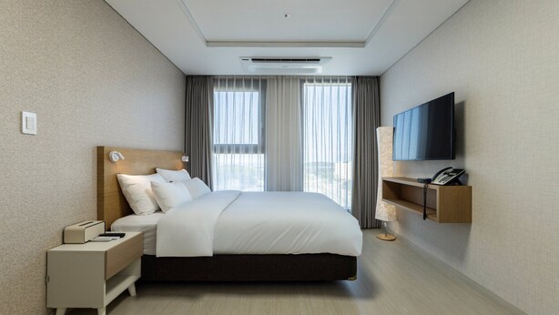 A modern hotel room with a large bed, a bedside table with a lamp, a TV mounted on the wall, and a window with curtains. - Centum Mark Kimstay Yangyang (Yangyang)