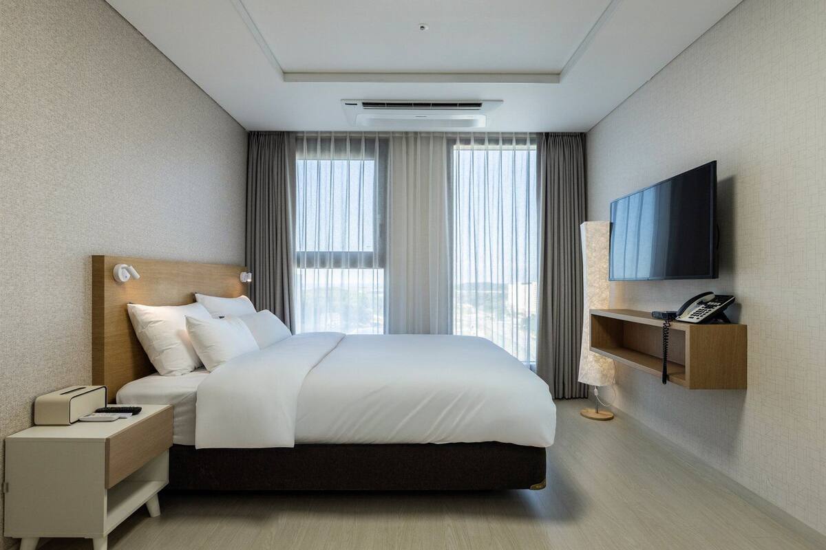 A modern hotel room with a large bed, a bedside table with a lamp, a TV mounted on the wall, and a window with curtains.