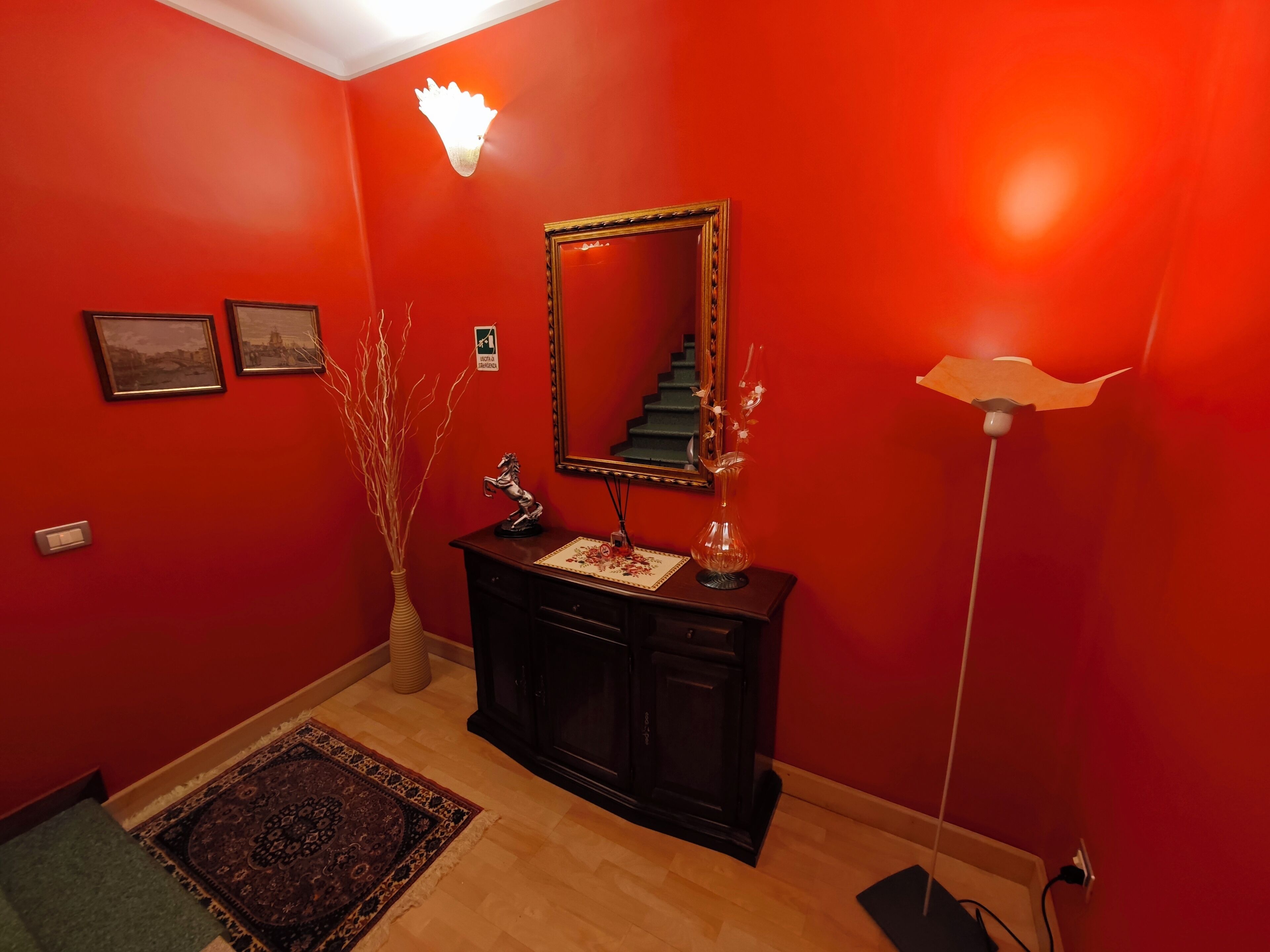 Una habitación con paredes rojas, piso de madera, una consola de madera oscura con un espejo, un florero y una pequeña estatua.