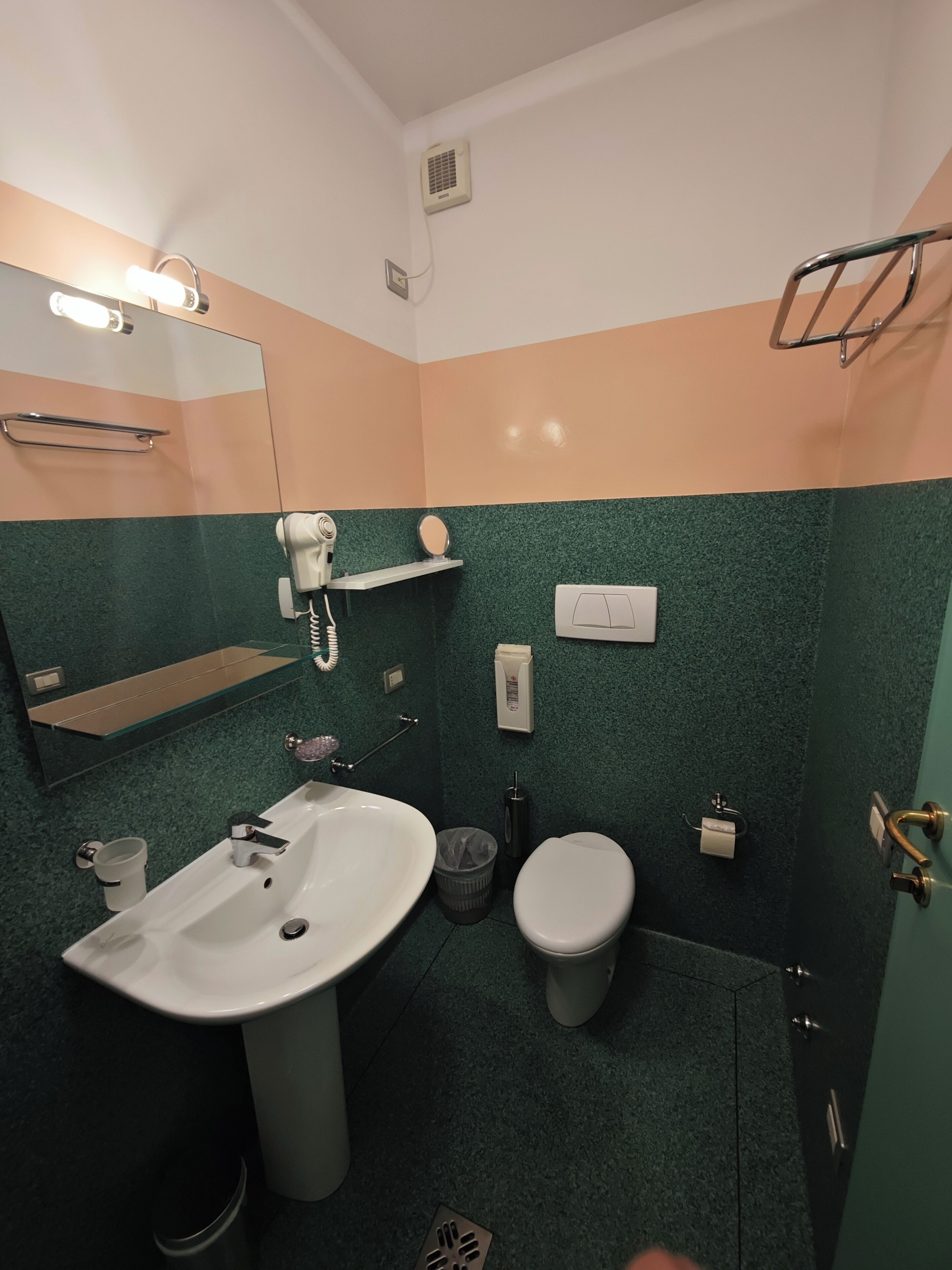 A bathroom with a sink, toilet, and mirror. There is a hairdryer on the wall, a towel rack, and a trash bin.