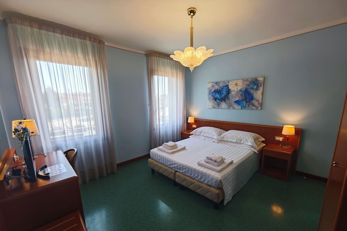 Habitación de hotel con cama, dos mesitas de noche con lámparas, un escritorio con lámpara, una silla y un cuadro en la pared.
