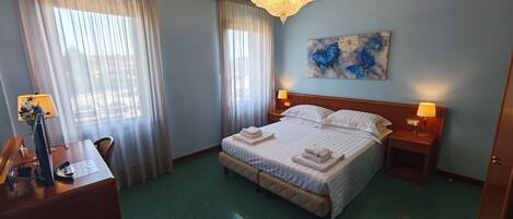 Habitación de hotel con cama, dos mesitas de noche con lámparas, un escritorio con lámpara, una silla y un cuadro en la pared.