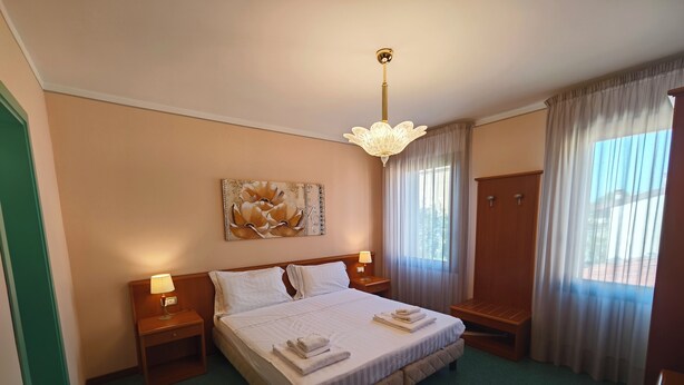 Quarto de hotel com uma cama grande, duas mesas de cabeceira com luminárias, um guarda-roupa de madeira, um tapete verde e uma pintura na parede.