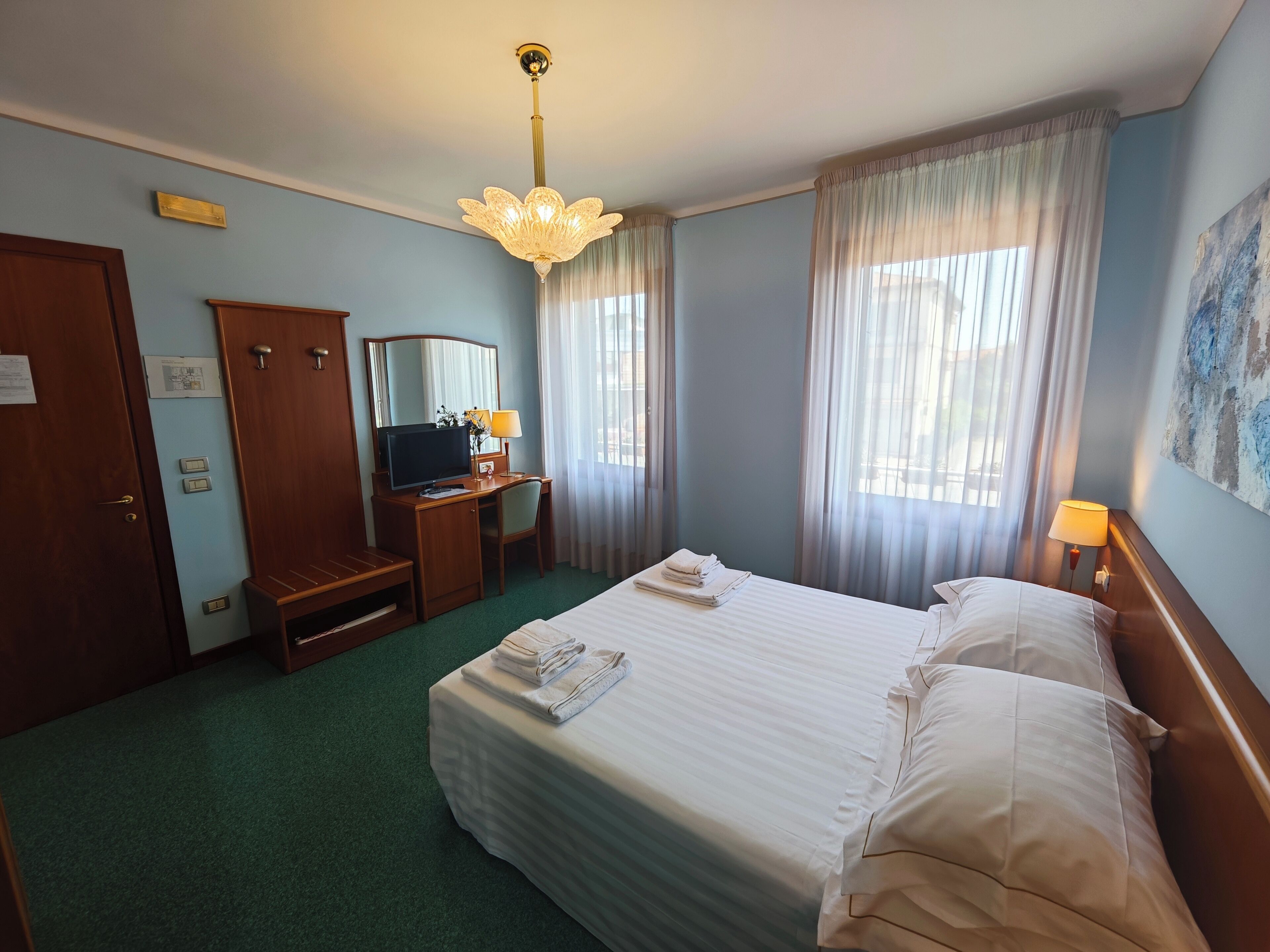 A hotel room with a bed, a desk with a computer, a television, a mirror, and a window with curtains.