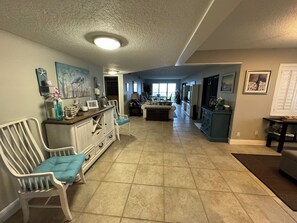 A spacious living area with a kitchen in the background, featuring a dining table, chairs, and a large window. - Spacious 2/2 Oceanfront Corner Unit with Incredible Direct Beach Views & Access! (Jensen Beach)