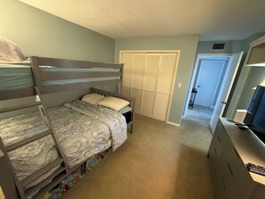 A bedroom with bunk beds, a closet with louvered doors, a desk with a lamp, and a door leading to another room. - Spacious 2/2 Oceanfront Corner Unit with Incredible Direct Beach Views & Access! (Jensen Beach)