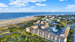 An aerial view of a coastal resort with beachfront properties, a swimming pool, and a lighthouse in the background.