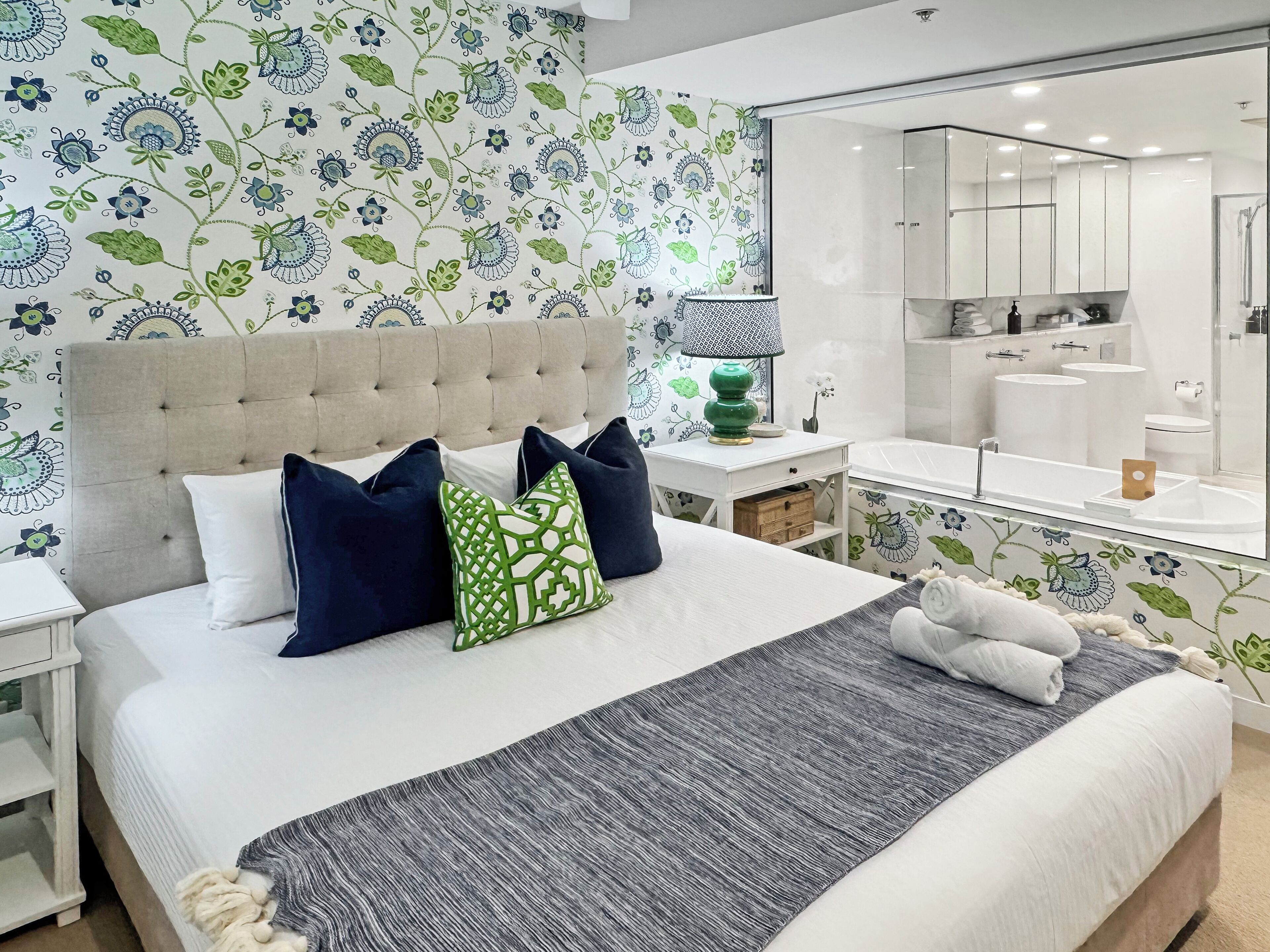 A bedroom with a large bed, a bedside table with a lamp, a nightstand, and a bathroom visible through an open door.