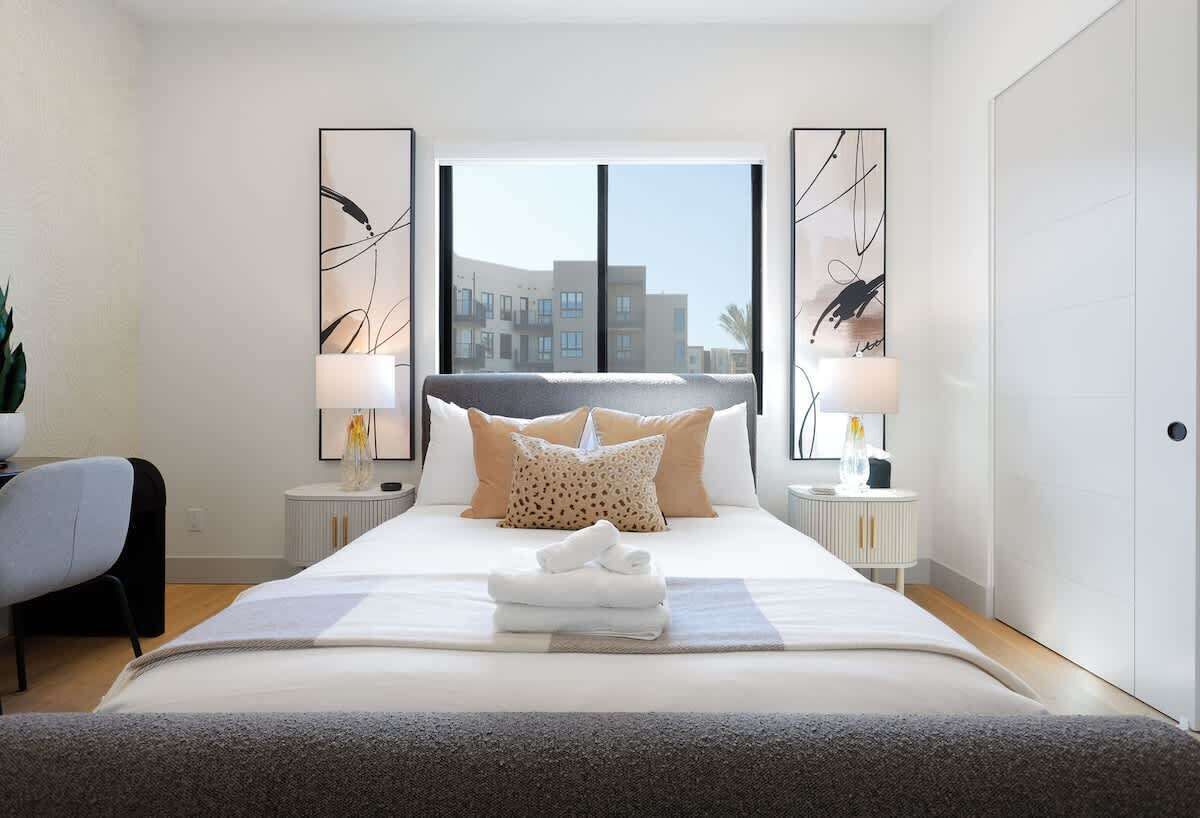 Habitación de hotel moderna con una cama grande, dos mesitas de noche con lámparas, vista a edificios por la ventana y arte abstracto en la pared.