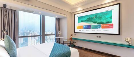 A hotel room with a large bed, a TV mounted on the wall, a view of the city through the window, and a small table with a vase of flowers.