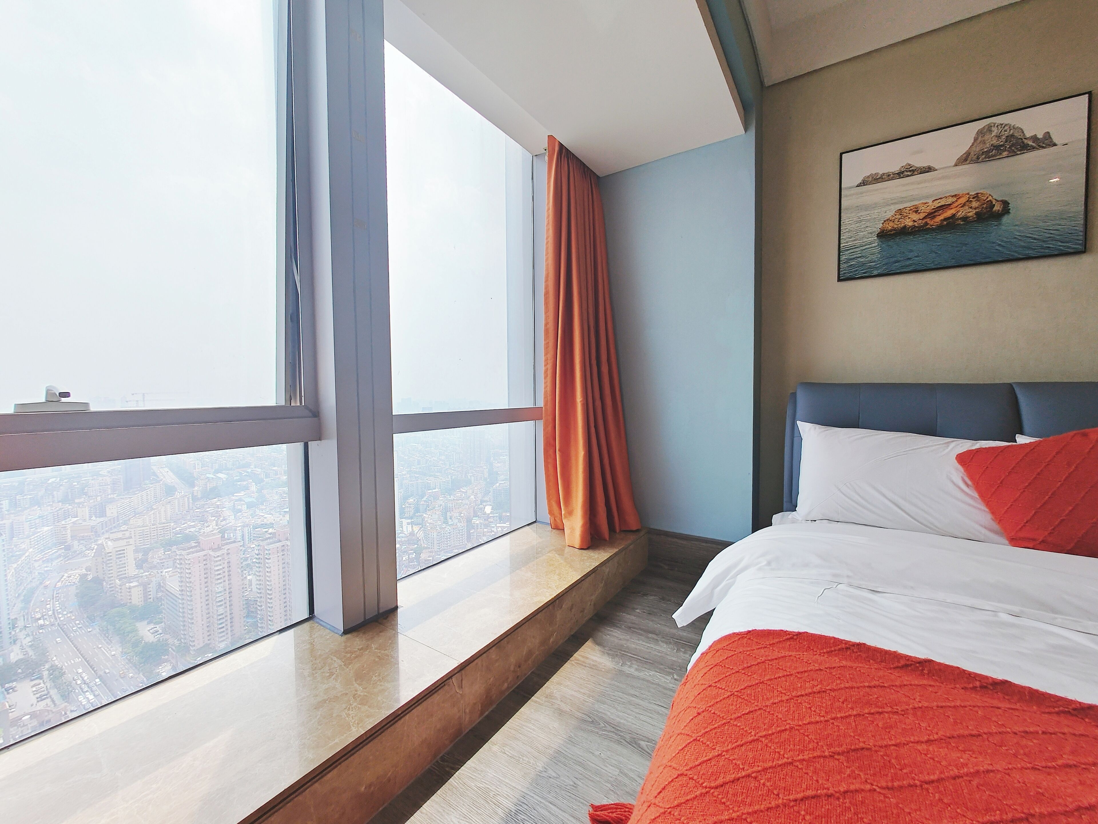 A hotel room with a large window offering a city view, a bed with white and red bedding, and a painting of a rocky coastline on the wall.