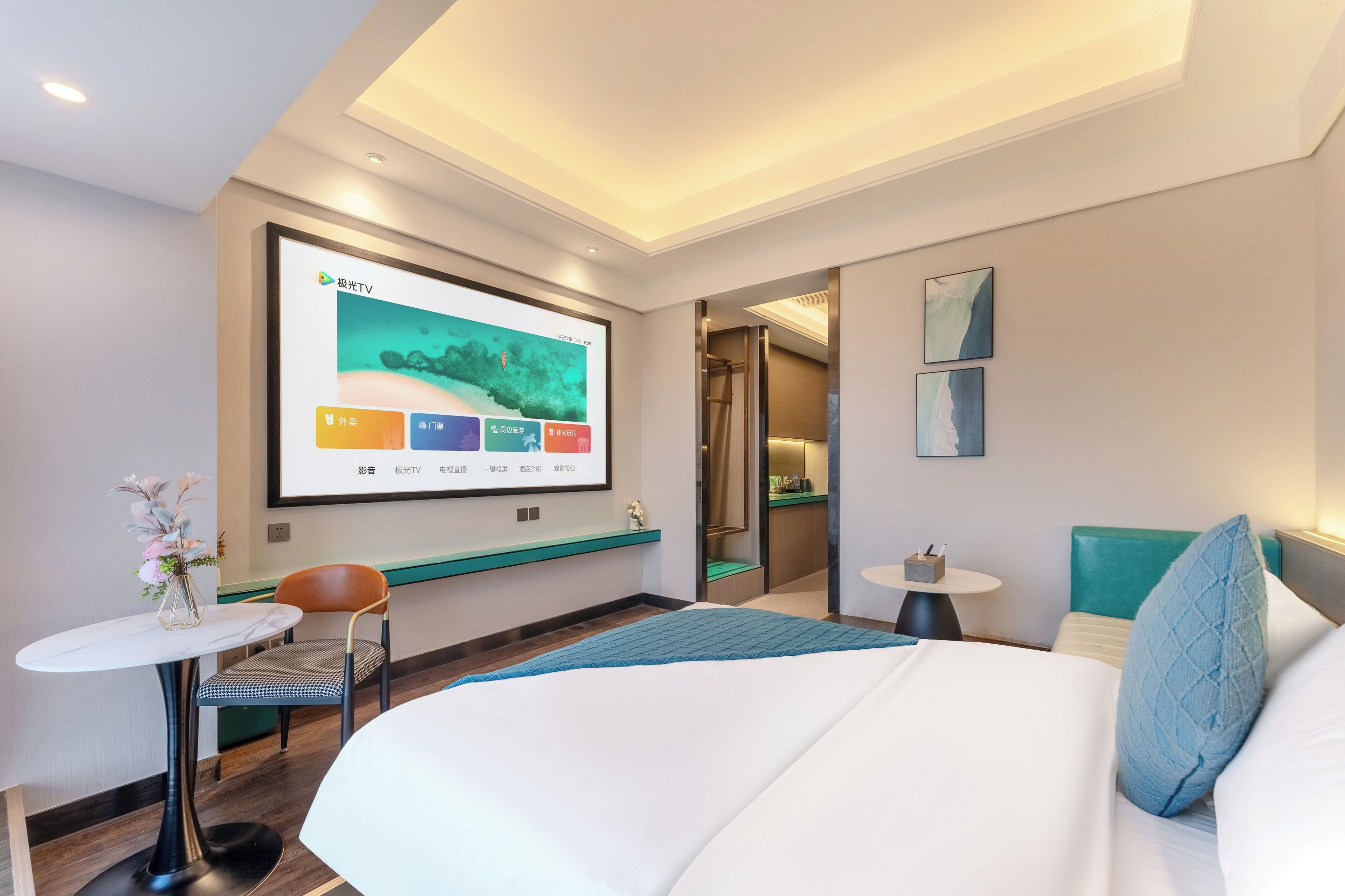 A hotel room with a bed, a desk, a chair, a small table, and a TV mounted on the wall displaying a weather forecast.