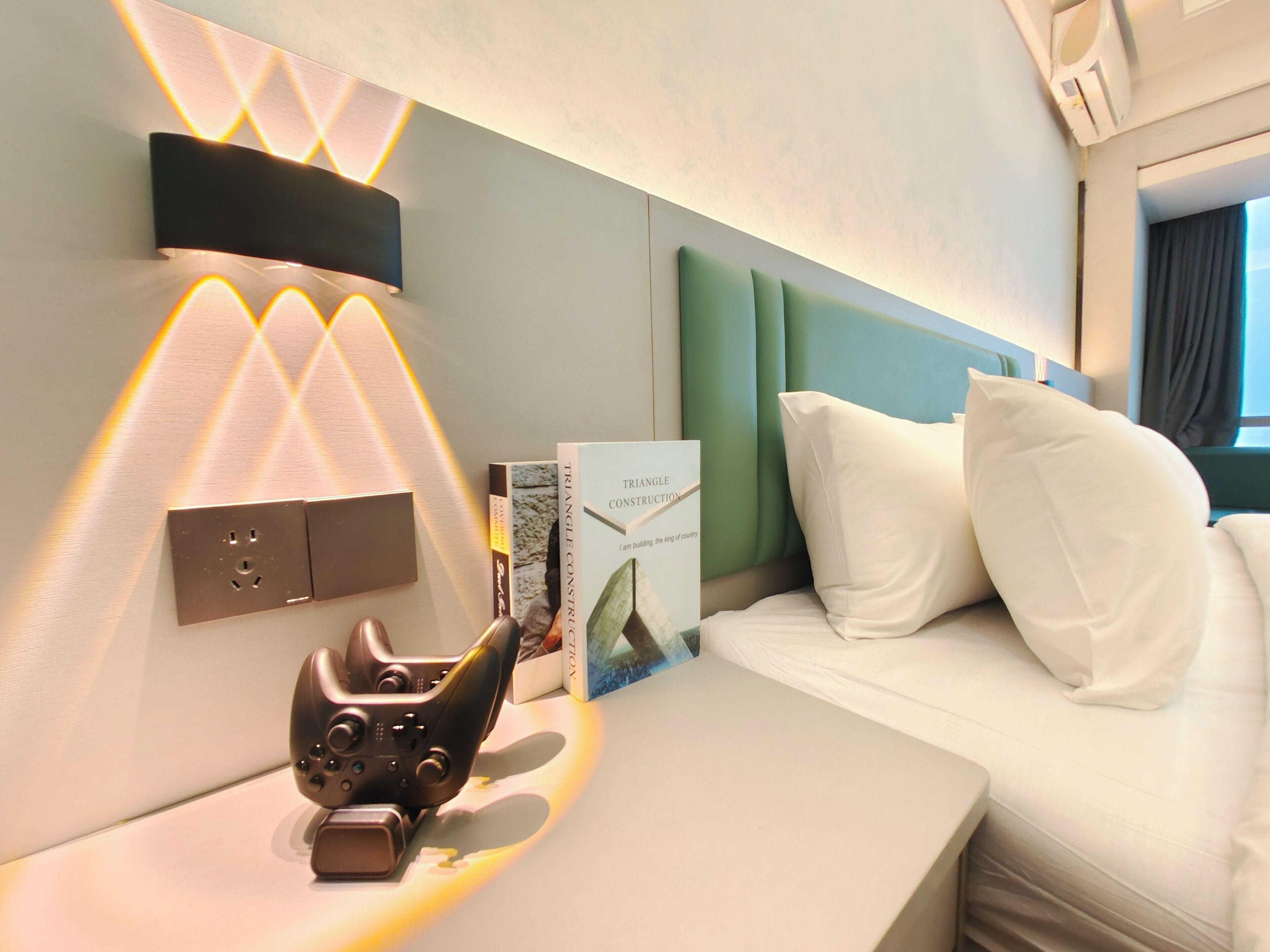 A hotel room with a bed, a bedside table with a game controller, a book titled 'Triangle Construction,' and a wall-mounted light fixture.
