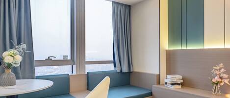 A modern hotel room with a seating area, a small table with a vase of flowers, and a bookshelf with books.