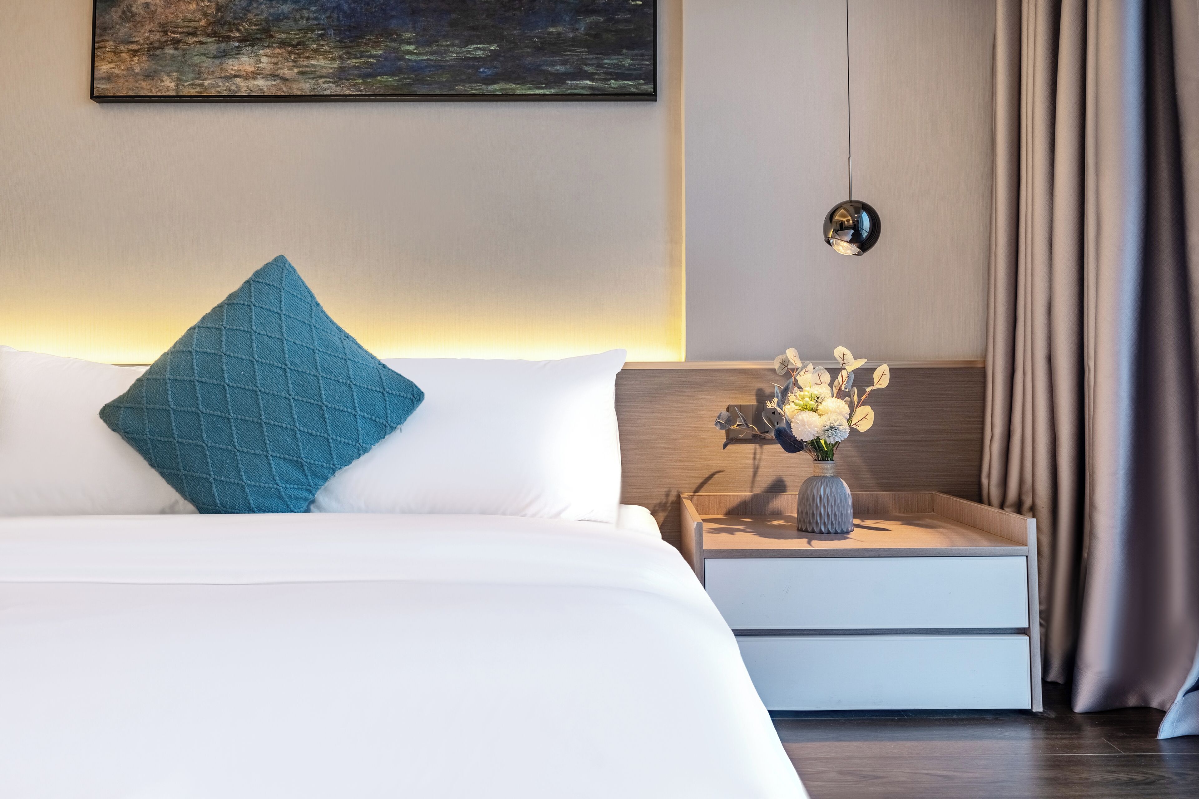 A modern hotel room with a neatly made bed, a bedside table with a vase of flowers, and a painting on the wall.