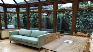 Une véranda avec une table et des chaises en bois, un canapé vert clair et de grandes fenêtres donnant sur un jardin.
