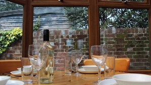 Une table à manger est dressée avec des verres à vin, une bouteille et des assiettes. On aperçoit, à travers la fenêtre, un mur de briques et de la verdure.
