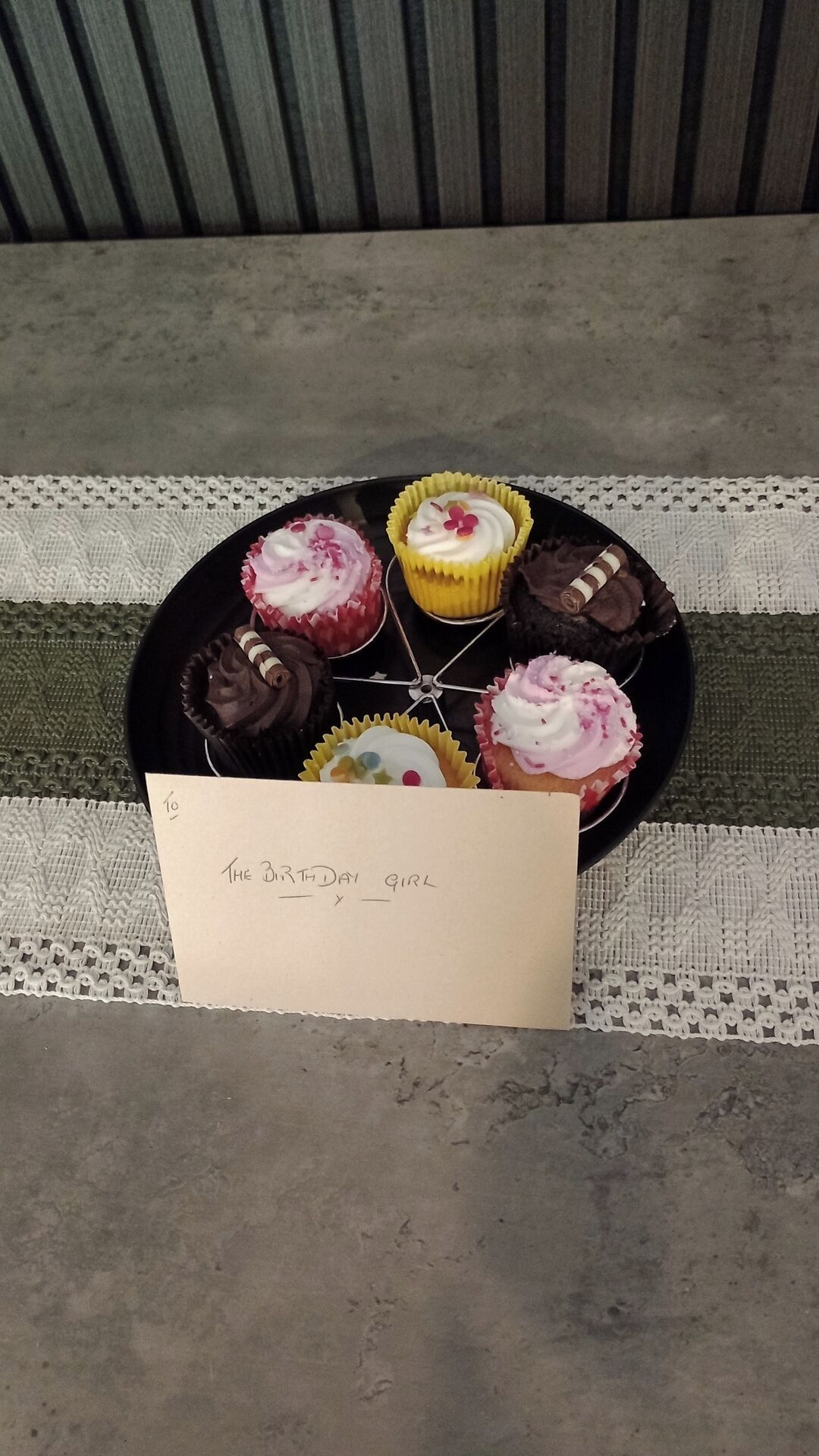 Une assiette noire sur laquelle sont disposés six cupcakes, le tout posé sur une nappe en dentelle. Une note manuscrite sur la table porte l’inscription : « À l’attention de l’invitée d’honneur. »