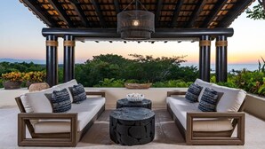 An outdoor seating area with two sofas and a round table, under a pergola with a hanging light fixture, overlooking a scenic view.