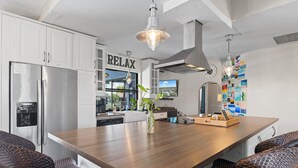 Una cocina moderna con armarios blancos, una isla de madera, sillas de mimbre y un cartel con la palabra 'RELAX'.